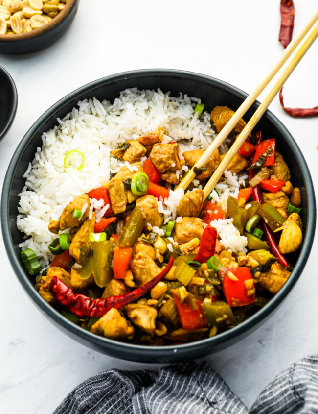 chinese chicken stir fry in a black bowl with rice and chopsticks.