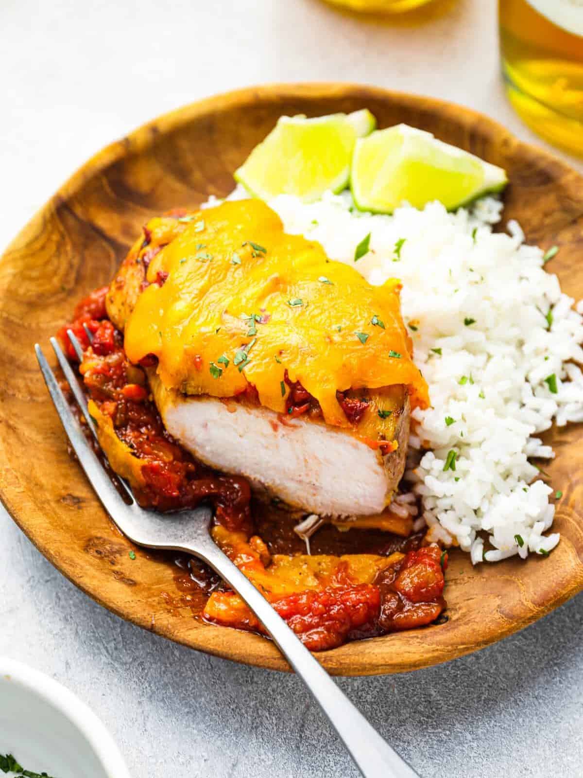 three-quarters view of a baked salsa chicken breast on a wooden plate with white rice, lime wedges, and a fork.