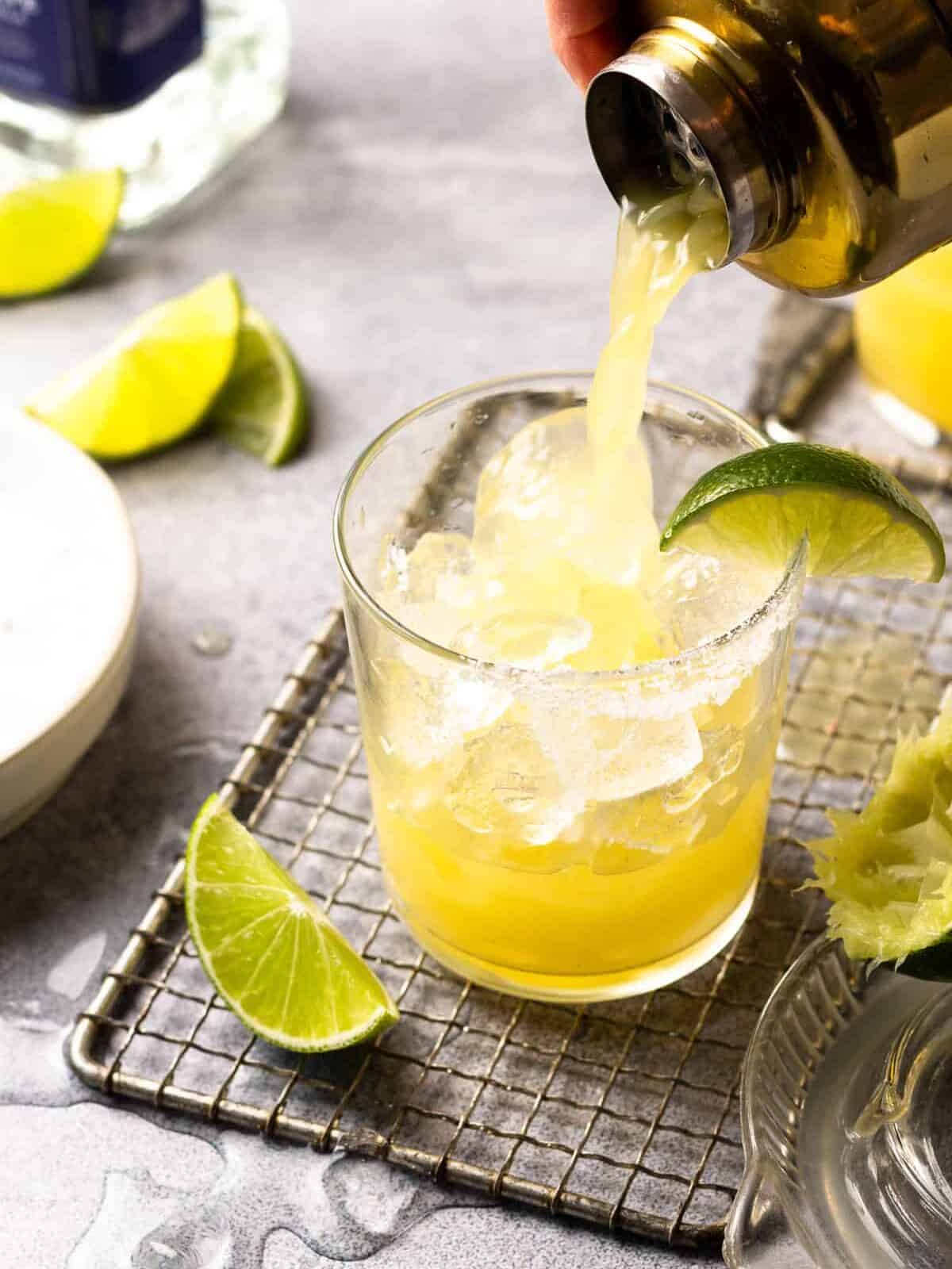 a cocktail shaker pouring a skinny margarita into a rocks glass filled with ice with a lime wedge on the rim.