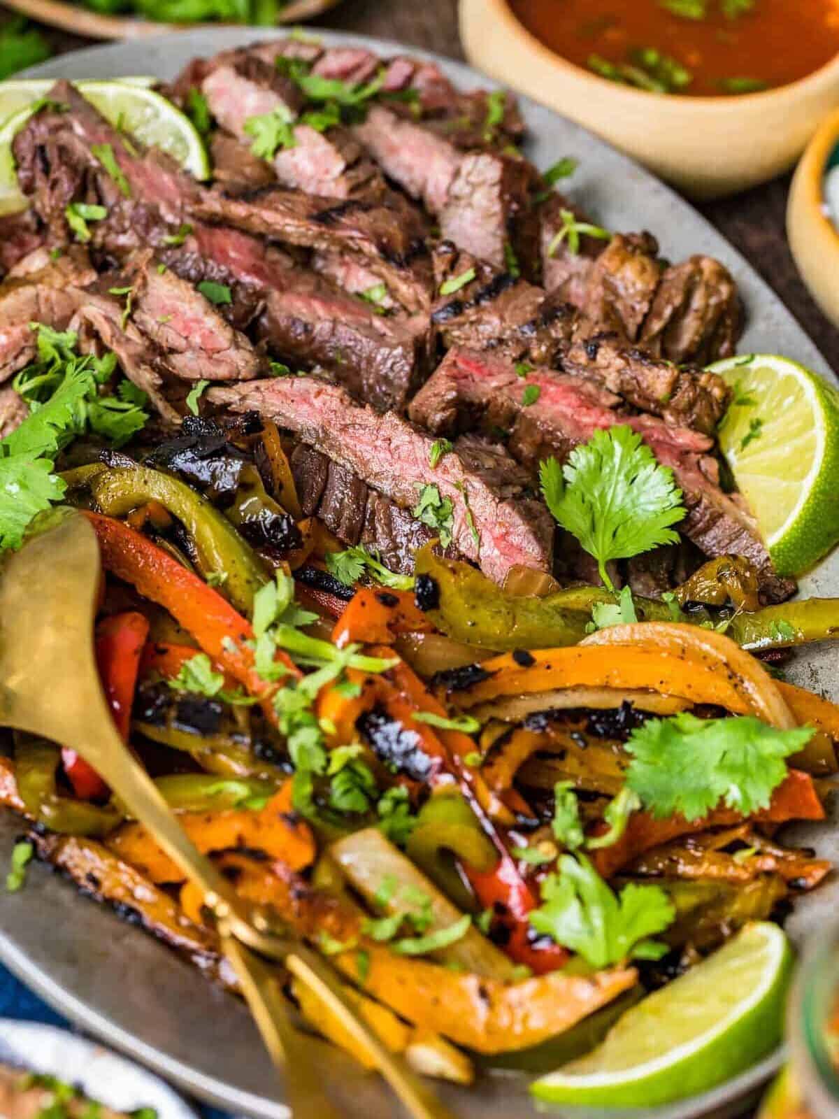 close up on a platter of marinated grilled steak and strips of fajita peppers and onions with a serving spoon