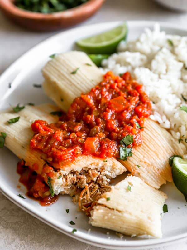 three-quarters view of 2 tamales on a white plate with cilantro rice and salsa, one tamale has been sliced into.