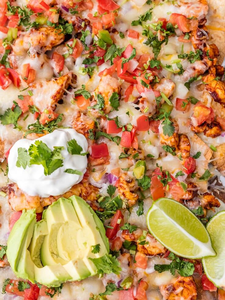 close up on baked chicken nachos topped with avocado slices, sour cream, lime wedges, pico de Gallo, and cheese