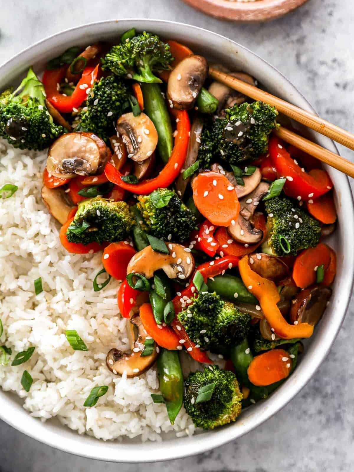 partial overhead view of vegetable stir fry in a white bowl with white rice and chopsticks.