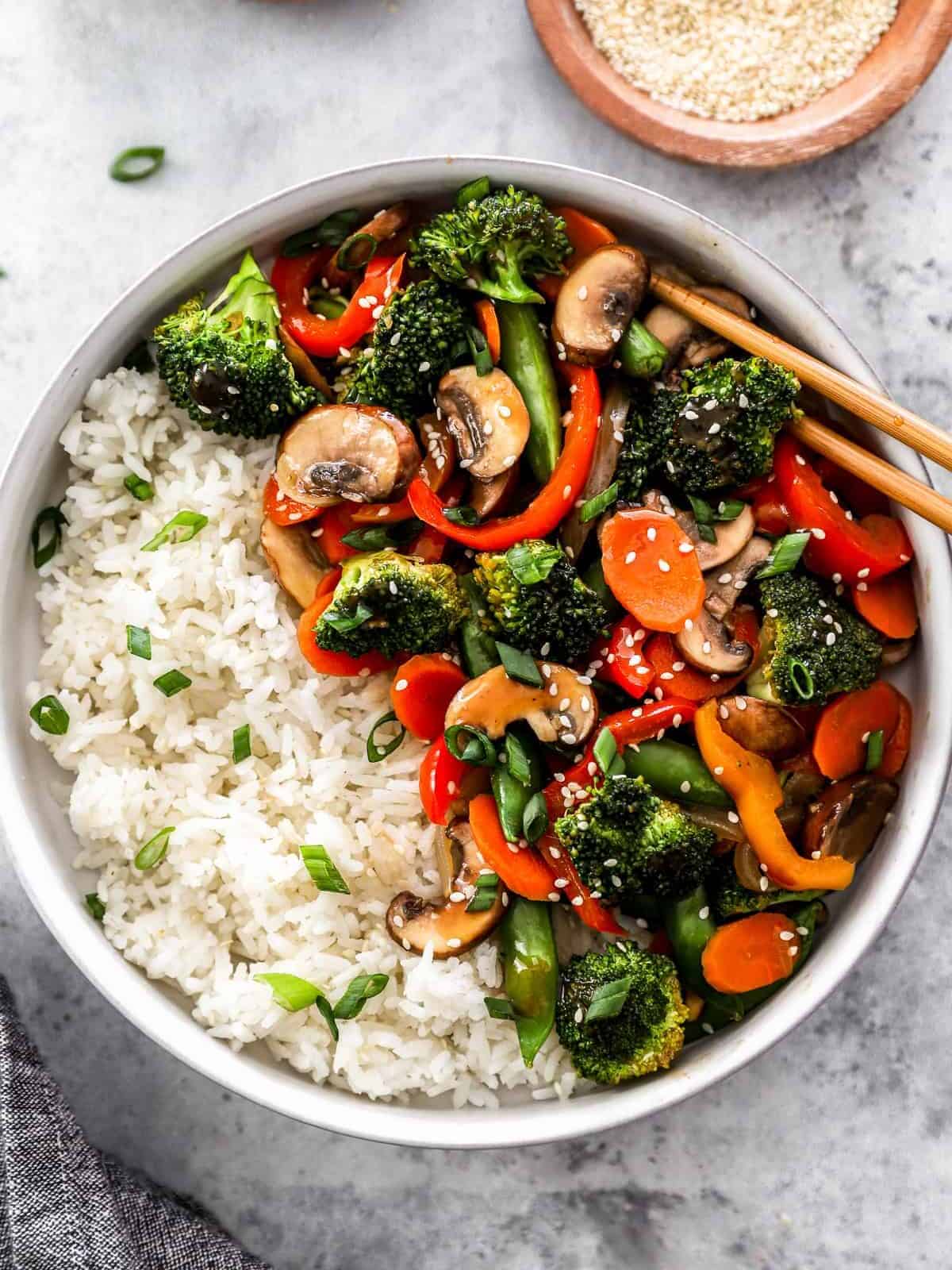 overhead view of healthy vegetable stir fry in a white bowl with white rice and chopsticks.