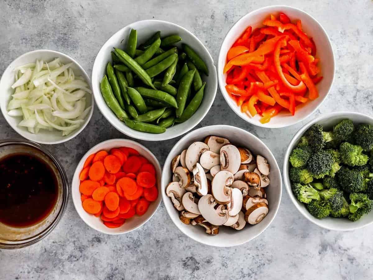 overhead view of stir-fried vegetables ingredients (broccoli, mushrooms, peppers, carrots, pea pods, onions) in individual bowls.