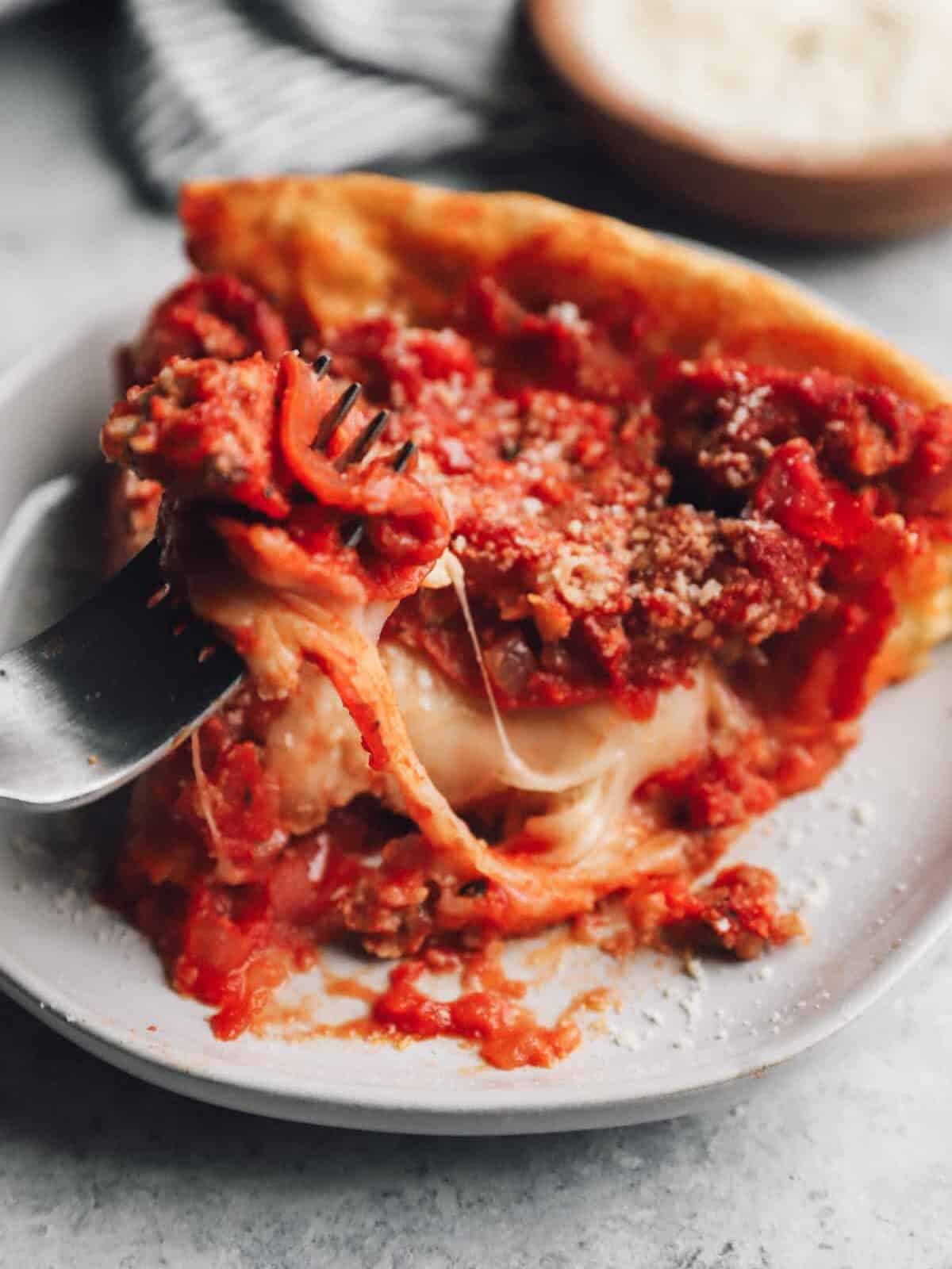 a fork removing a bite of chicago deep dish pizza from a slice on a white plate.