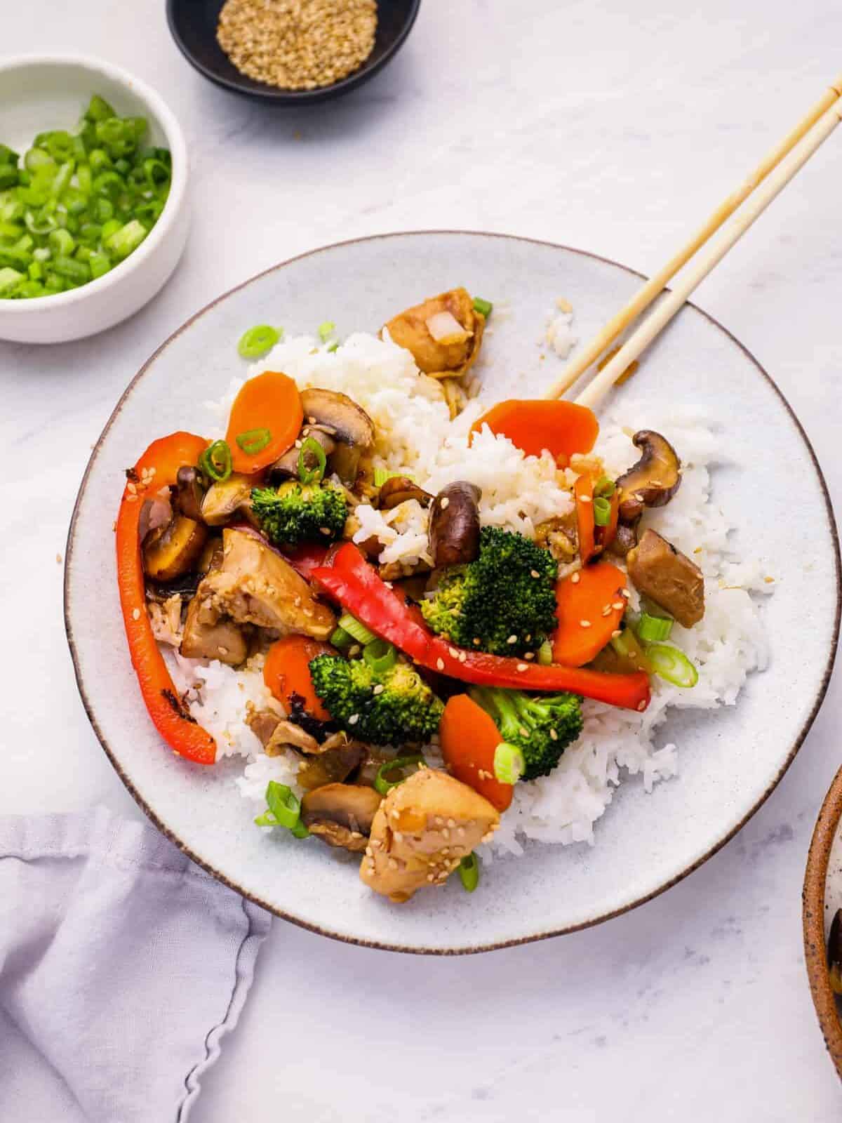 chicken stir fry over rice on a white plate with chopsticks.