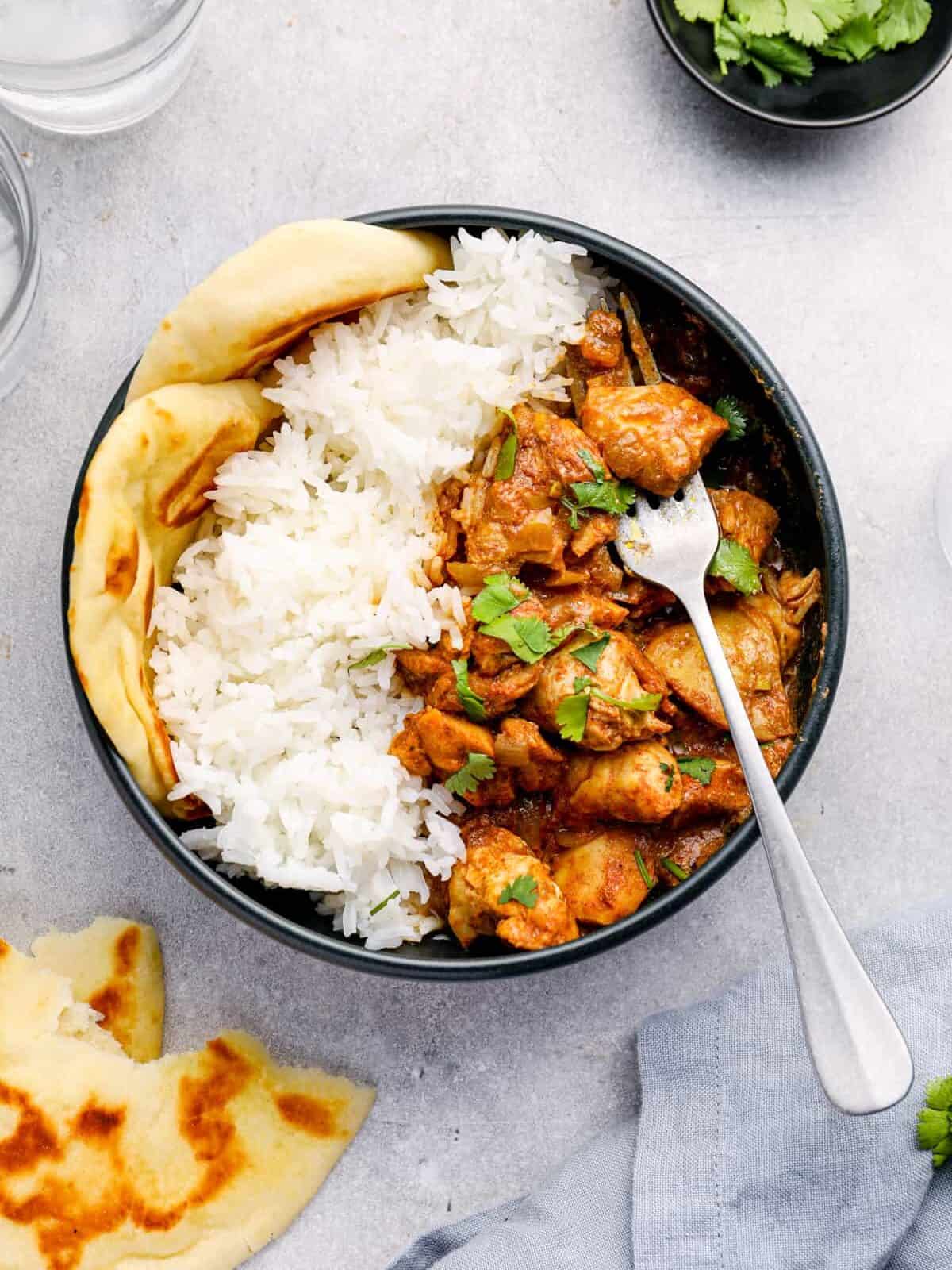 overhead view of crockpot chicken tikka masala with rice and naan in a black bowl with a fork.