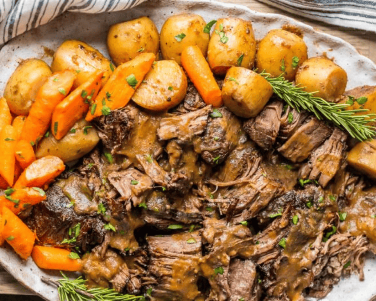 Shredded pot roast served on a white platter with tender carrots, baby potatoes, and rich brown gravy, garnished with fresh parsley and rosemary.