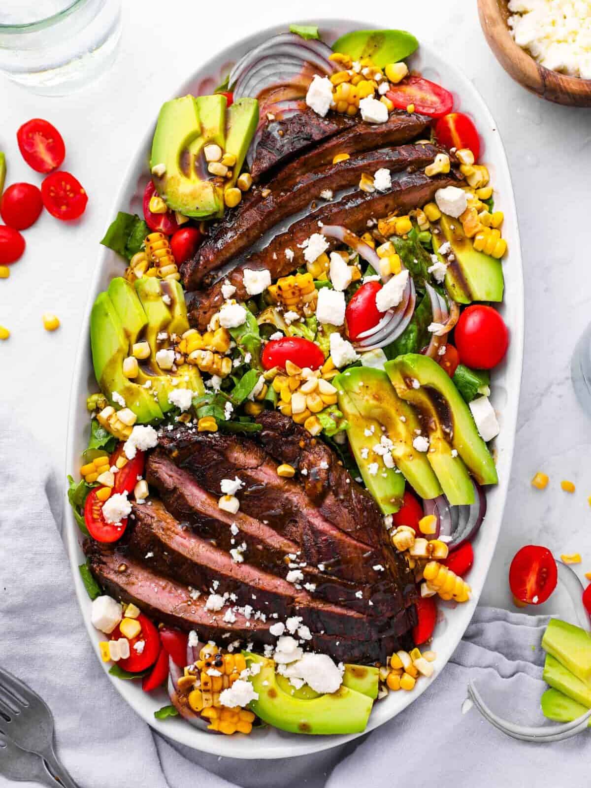 overhead view of salad with marinated steak, grilled corn, avocado, tomatoes, and more, in a white oval serving platter.