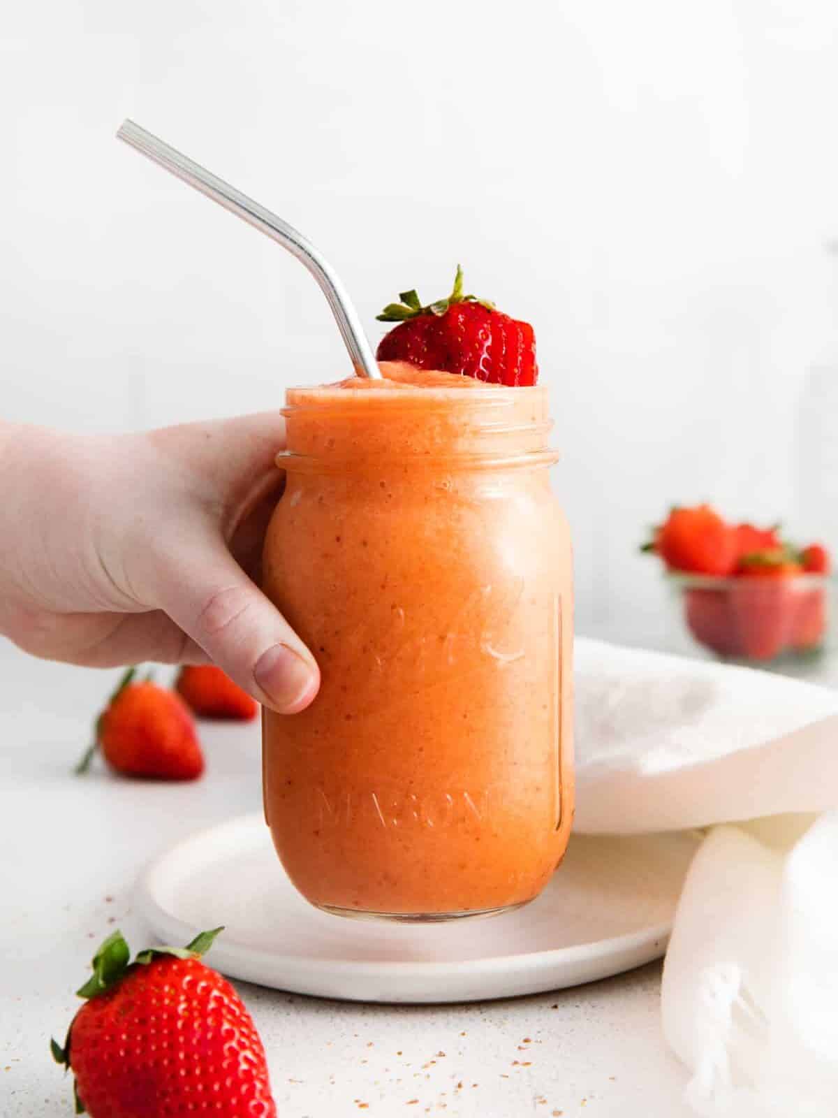 a hand grabbing a strawberry mango smoothie in a mason jar with a stainless steel straw.