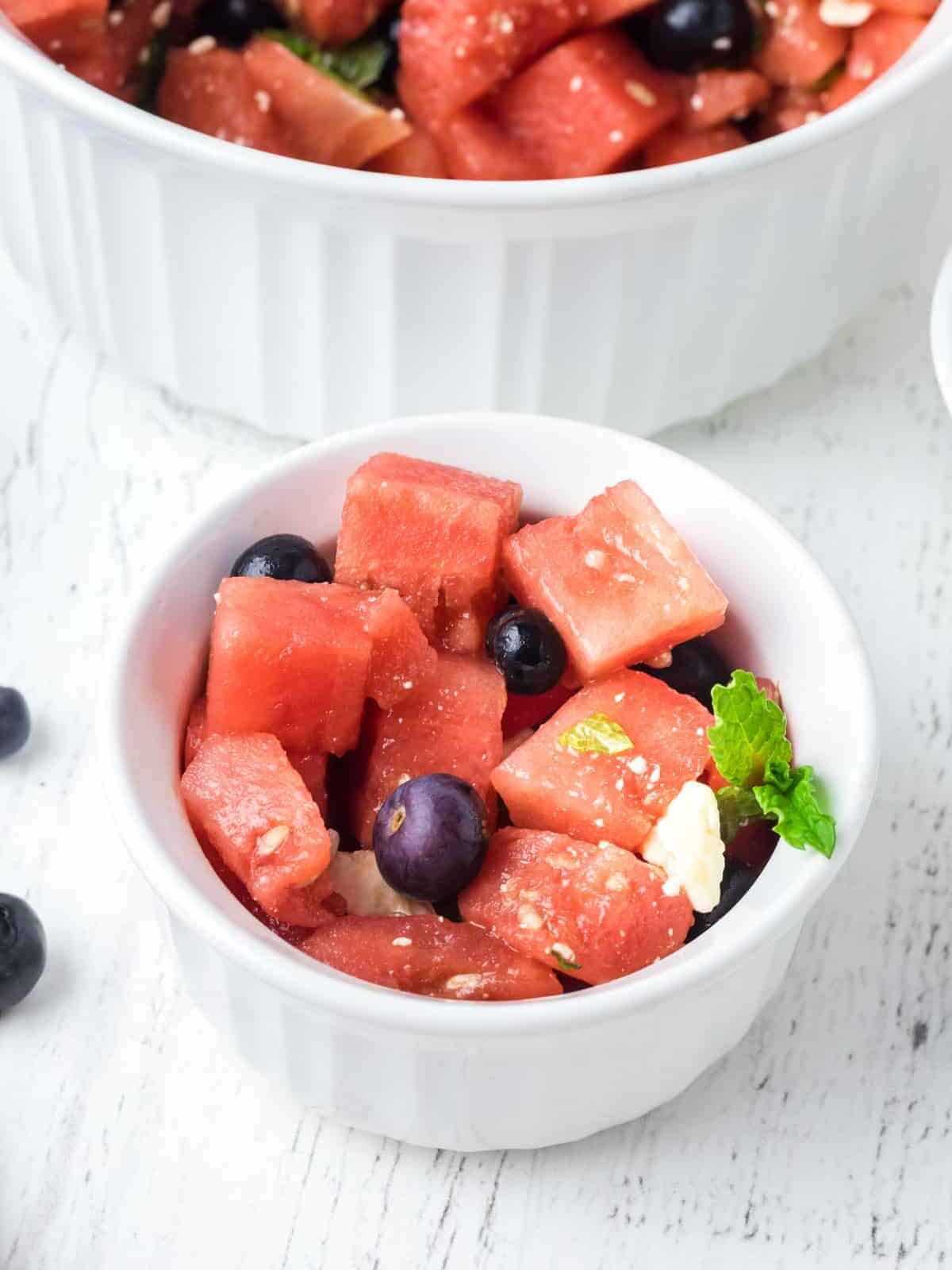 three-quarters view of a small bowl of watermelon salad in front of a large serving bowl.