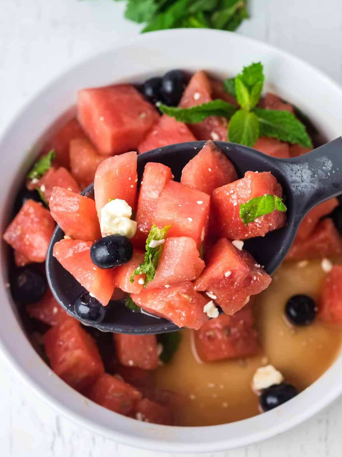 a spoon lifting watermelon salad from a white serving bowl.