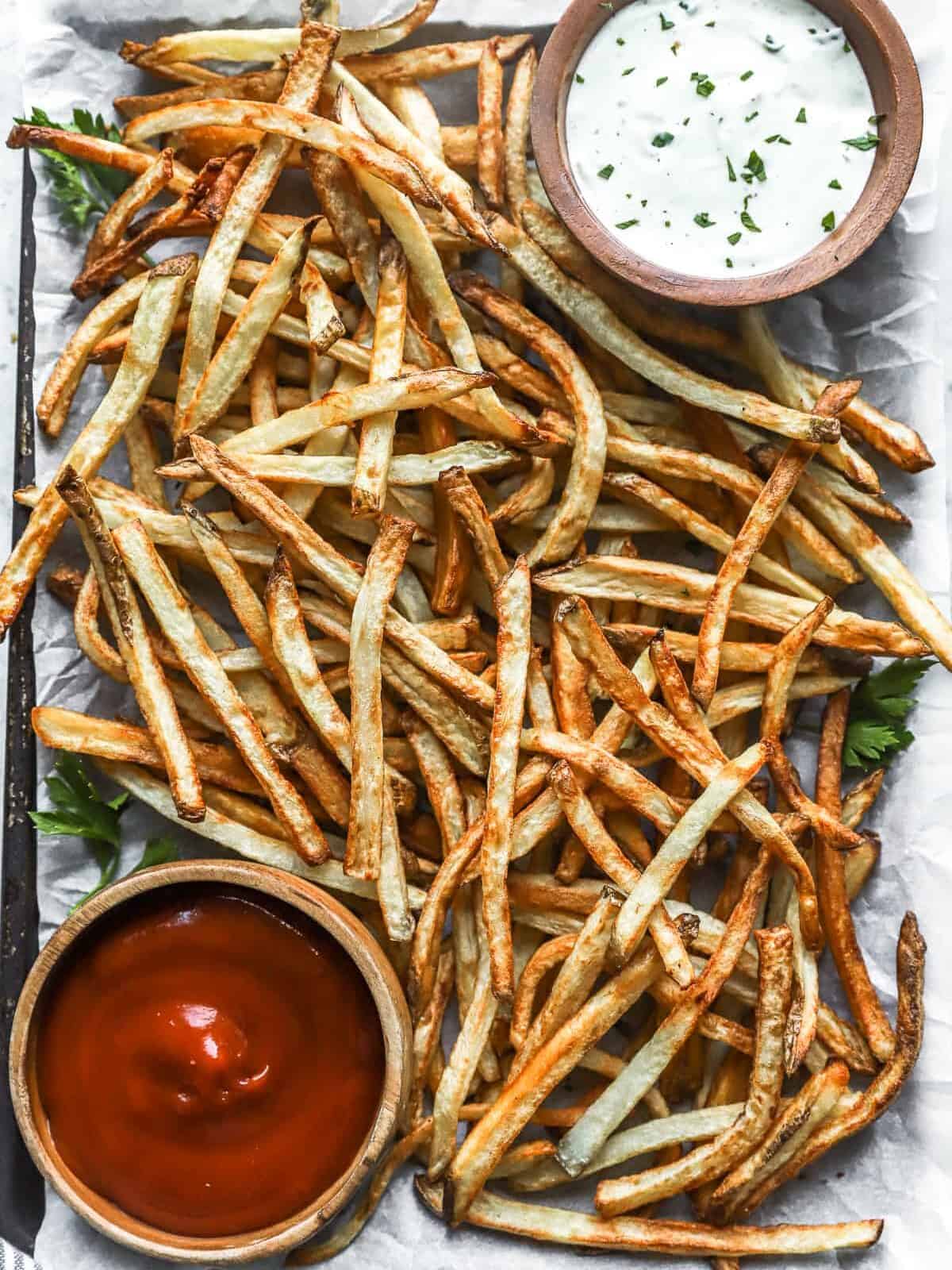 overhead view of air fryer french fries on a baking sheet with a cup of ketchup in the lower left corner and a cup of ranch in the upper right corner.