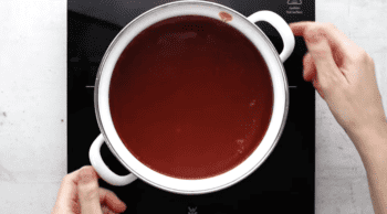 A person preparing a Bloody Mary cocktail by pouring red liquid into a pot on a stove.