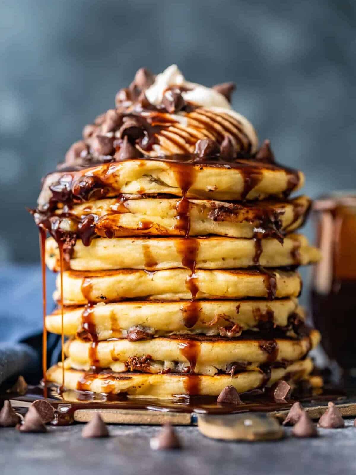 lots of chocolate chip pancakes stacked on each other drizzled with chocolate syrup and whipped cream