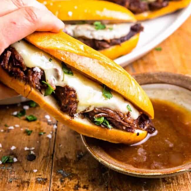 side view of a hand dipping a crockpot french dip sandwich into a side of au jus.