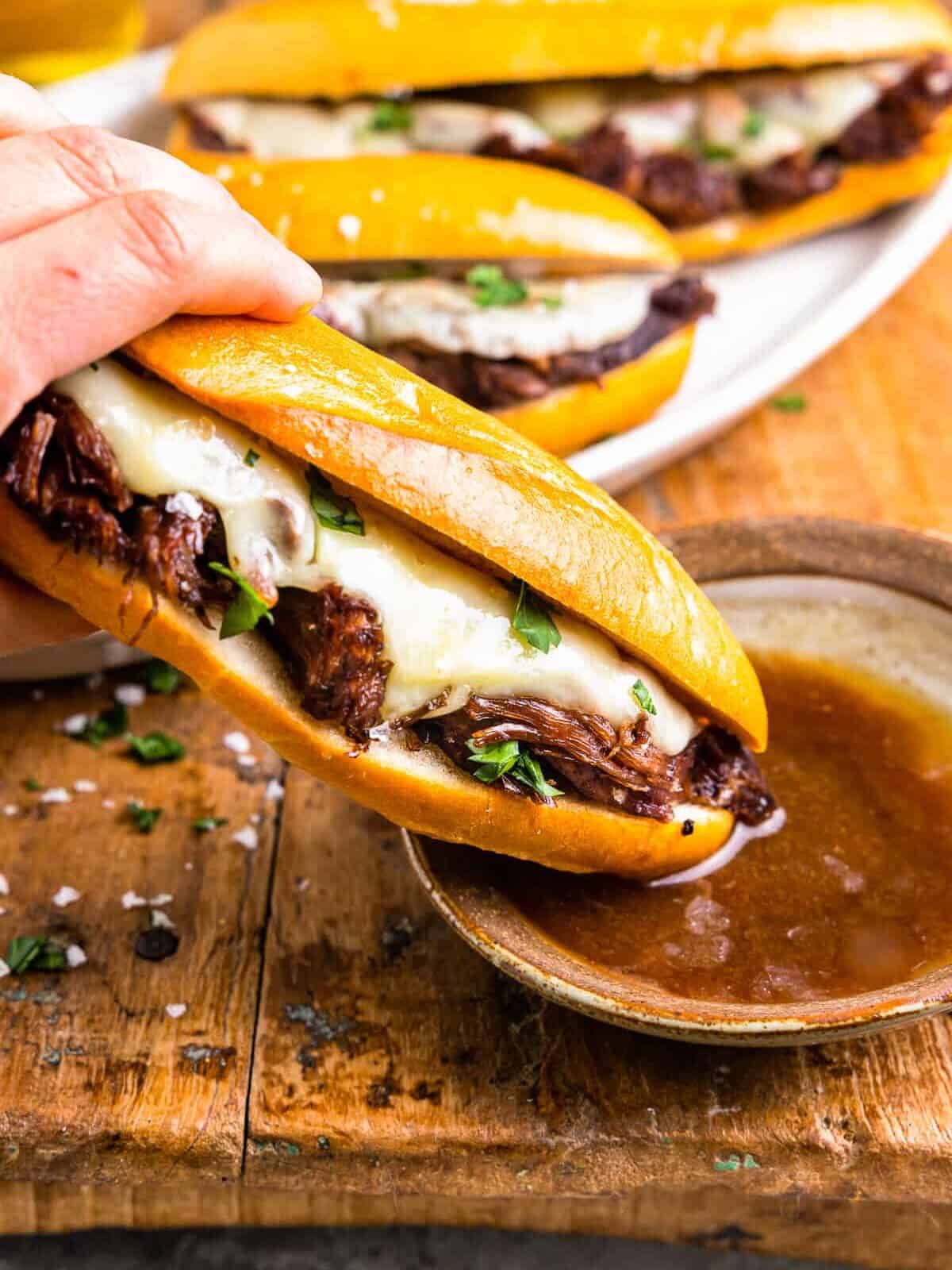 side view of a hand dipping a crockpot french dip sandwich into a side of au jus.