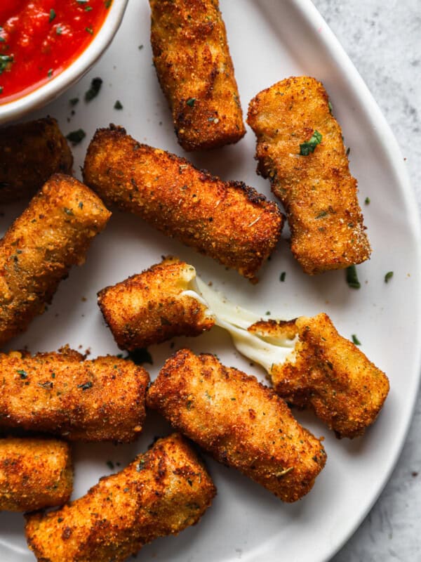 close up overhead view of mozzarella sticks on a white serving platter with a bowl of marinara sauce, one is halved.