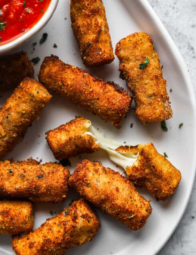 close up overhead view of mozzarella sticks on a white serving platter with a bowl of marinara sauce, one is halved.