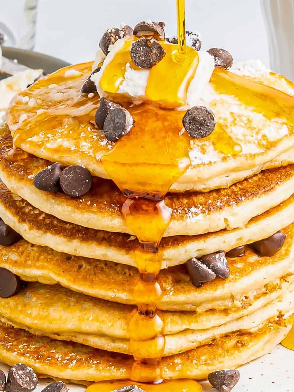 a tall stack of gluten free pancakes on a white plate with chocolate chips maple syrup and butter.