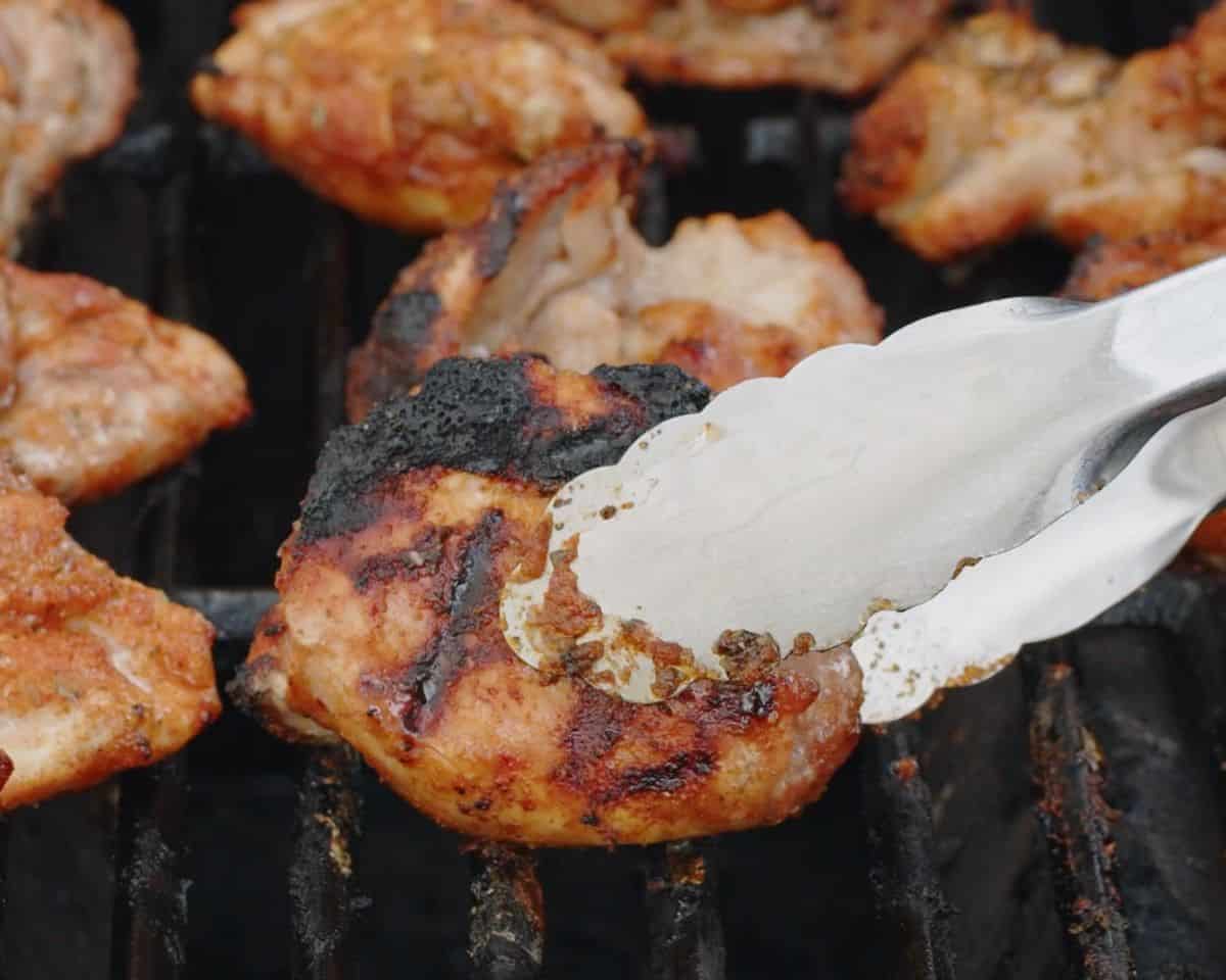 Close-up of grilled chicken thigh being turned with metal tongs on a hot grill, showing charred edges and caramelized seasoning.