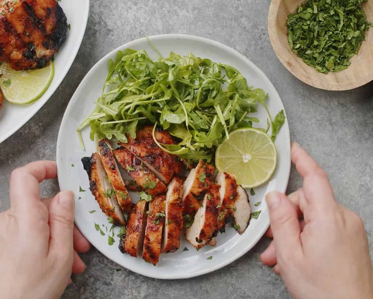 Sliced grilled chicken thighs served with fresh arugula, lime wedges, and chopped cilantro on a white plate.