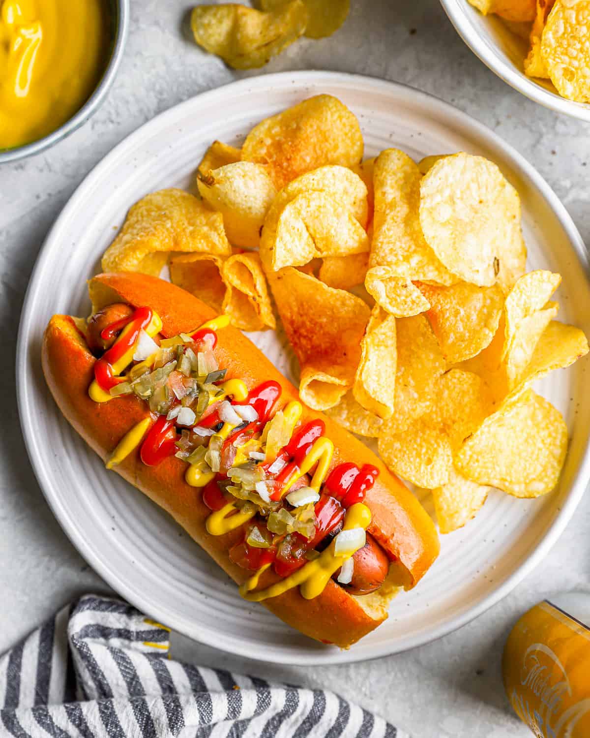 overhead view of a hot dog wit toppings on a white plate with potato chips.