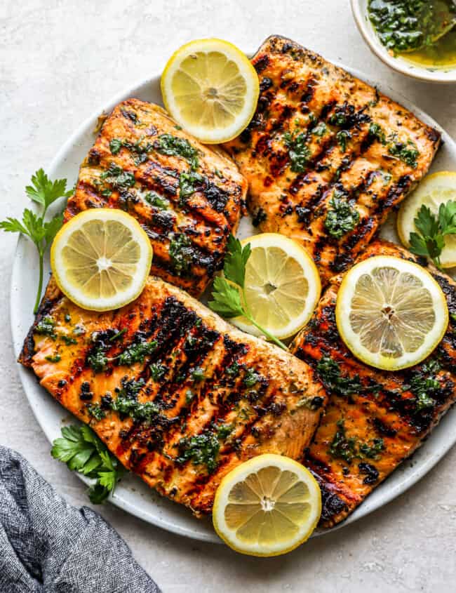 overhead view of 4 overlapping grilled salmon fillets on a white plate topped with herbs and lemon slices.