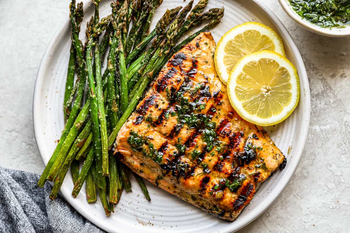 overhead view of a grilled salmon fillet on a white plate with asparagus and lemon slices.