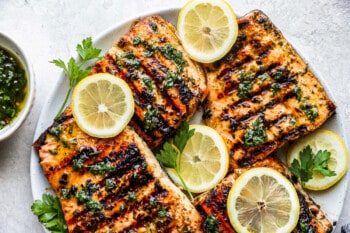 4 overlapping grilled salmon fillets on a white plate with herbs and lemon slices.