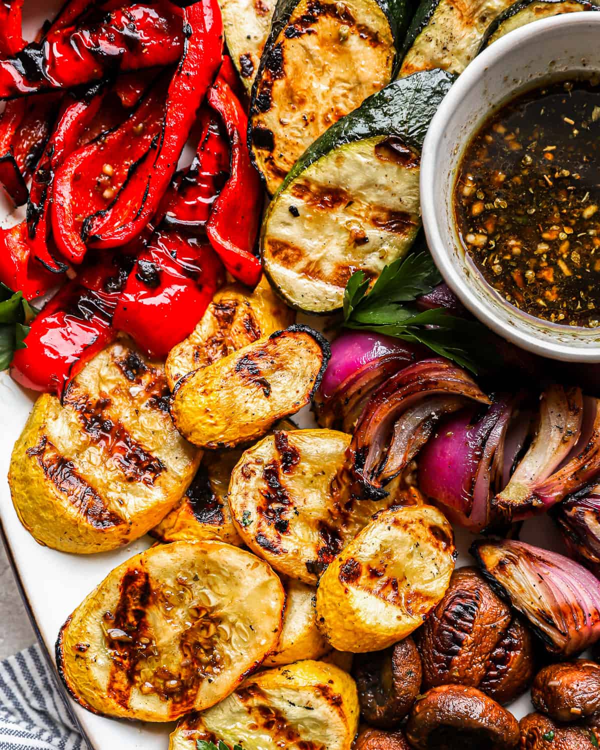 close-up overhead view of grilled vegetables on a white oval plate with marinade in a small bowl.