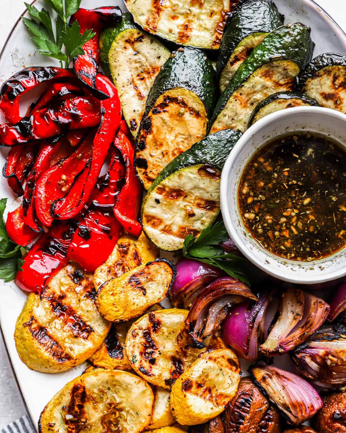 medium-distance overhead view of grilled vegetables on a white oval plate with marinade in a small bowl.