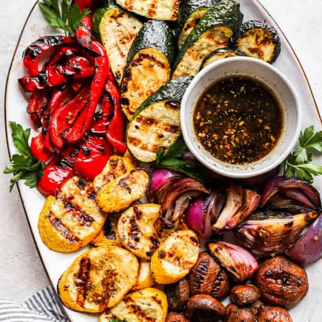 overhead view of grilled vegetables on a white oval plate with marinade in a small bowl.