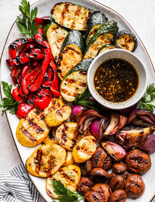 overhead view of grilled vegetables on a white oval plate with marinade in a small bowl.