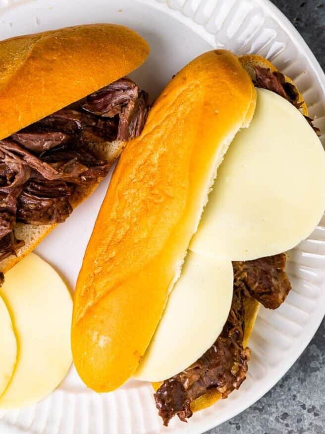 an assembled crockpot french dip sandwich on a white plate next to a crockpot french dip sandwich without any cheese.