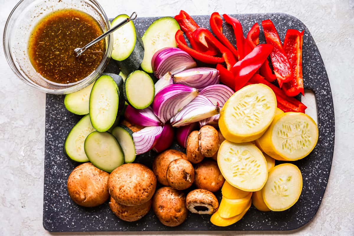 overhead view of ingredients for grilled vegetables on a cutting board. a mix of sliced veggies, including zucchini, summer squash, onions, bell peppers, and mushrooms are piled onto a cutting board.