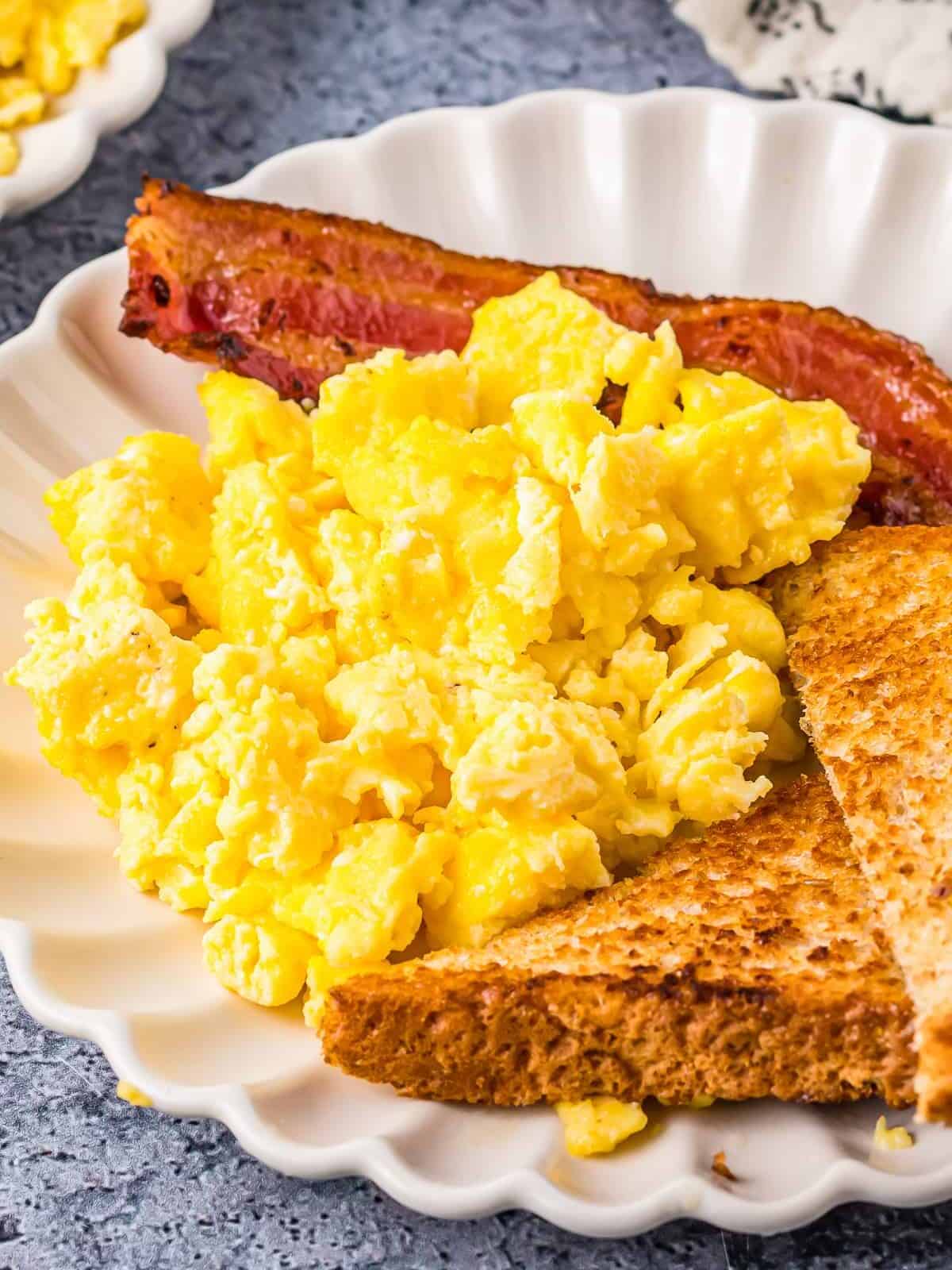 three-quarters view of scrambled eggs on a white plate with toast and bacon.