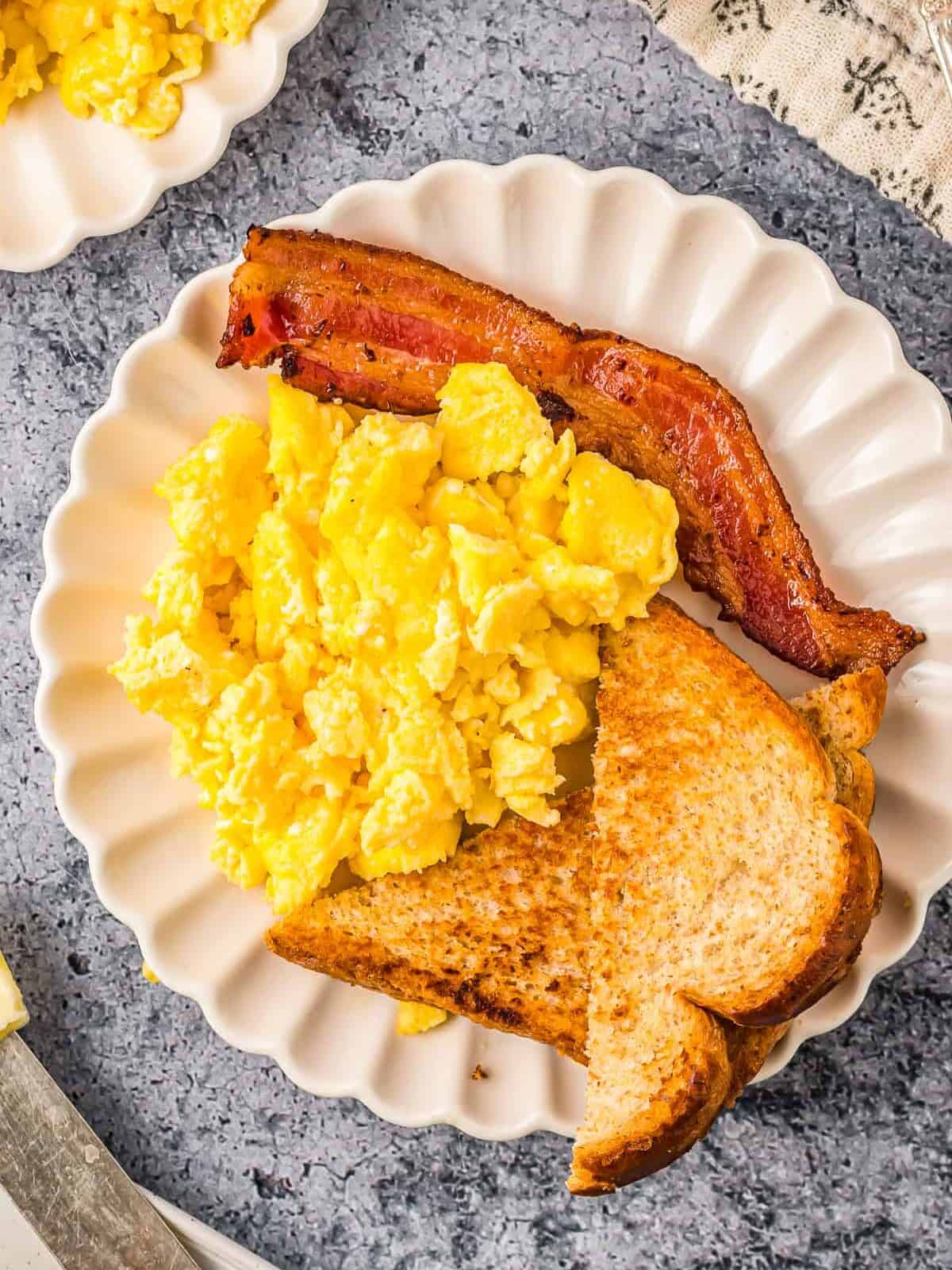 overhead view of scrambled eggs on a white plate with toast and bacon.