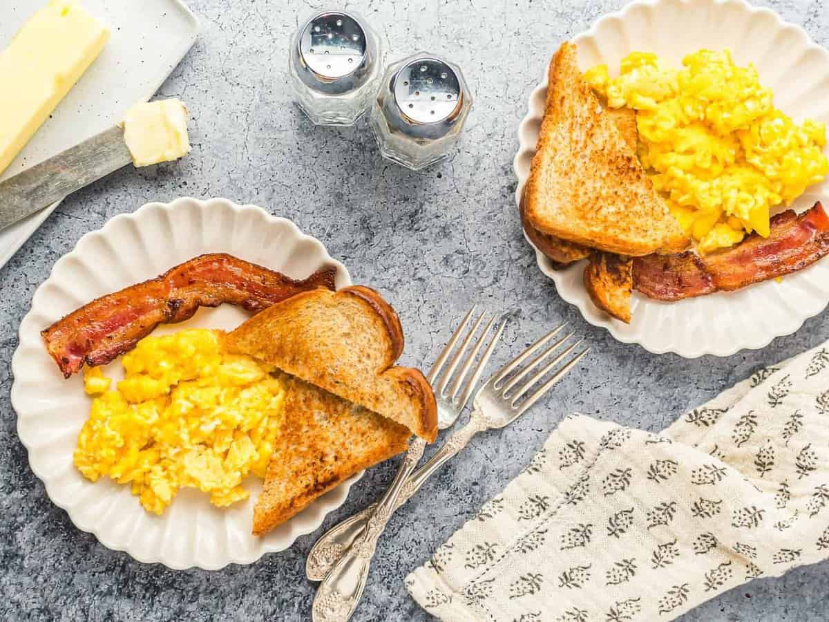 overhead view of 2 white plates with scrambled eggs, toast, and bacon on them.