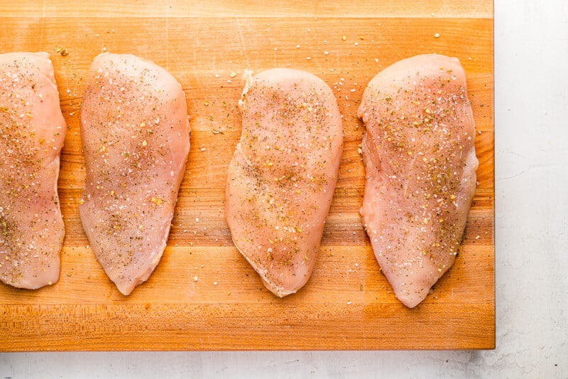 4 raw seasoned chicken breasts on a wooden cutting board.