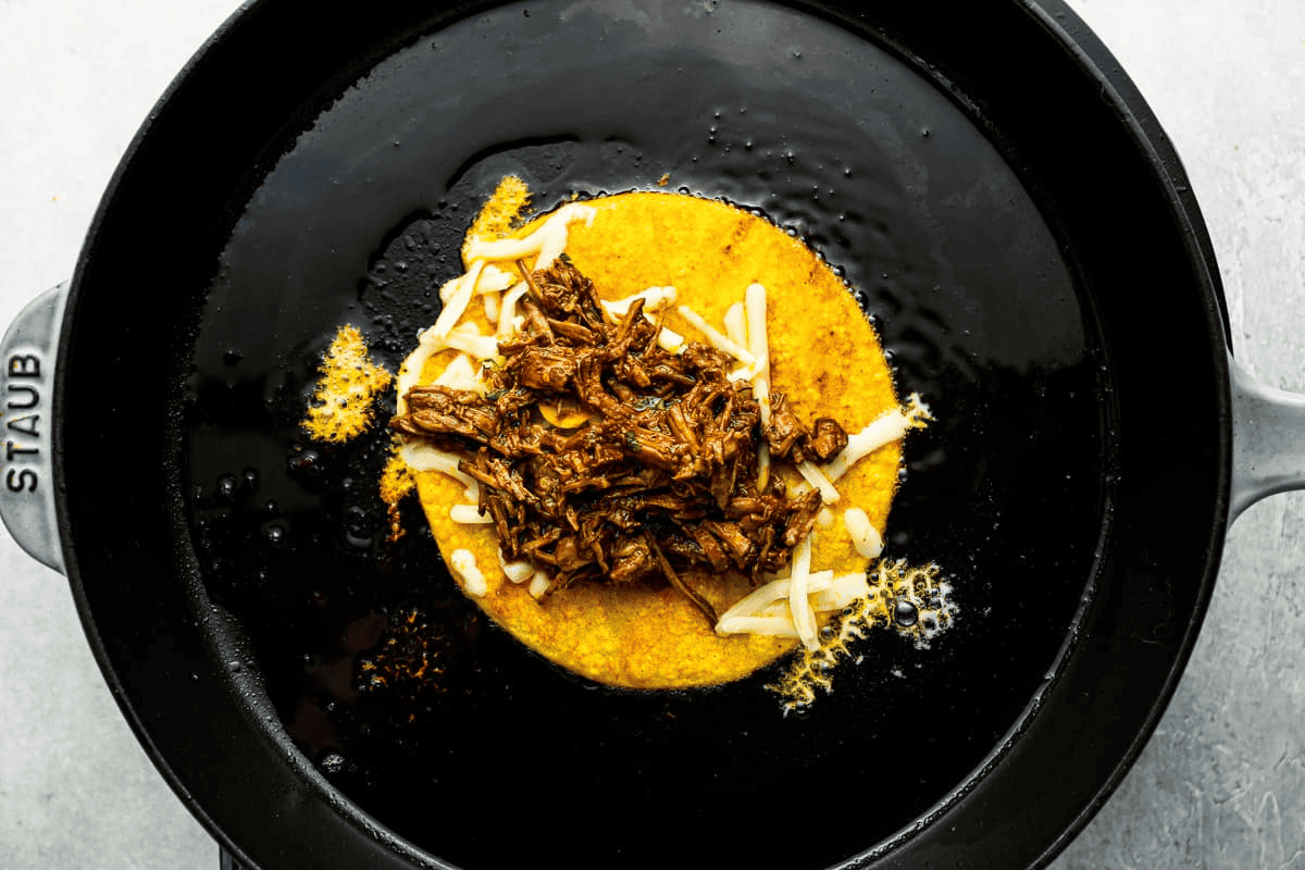 Cooking the quesabirria taco topped with meat and cheese, in a black cast iron pan.