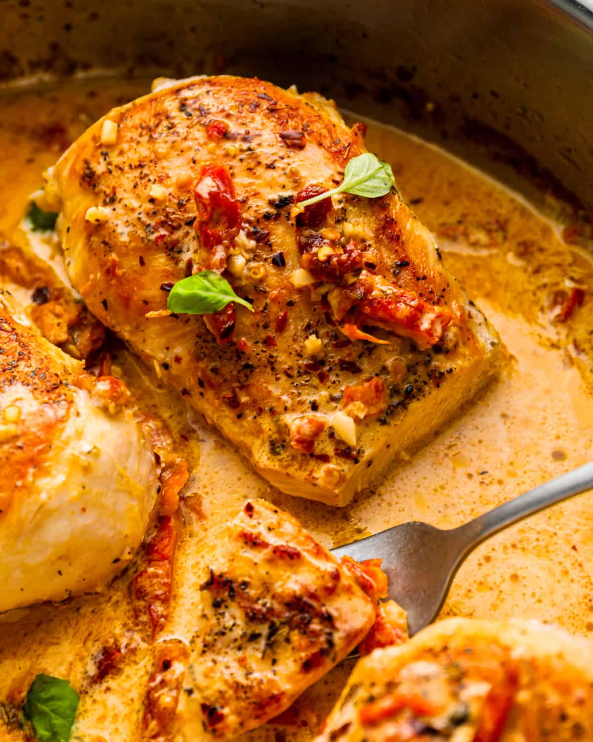 close up view of a forkful of marry me chicken with sun-dried tomatoes, next to a cut chicken breast.