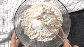 A person preparing a batch of peanut butter blossoms by mixing flour in a bowl with a whisk.