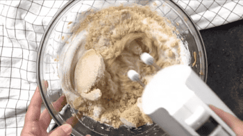 A person preparing peanut butter blossoms by mixing ingredients in a glass bowl.