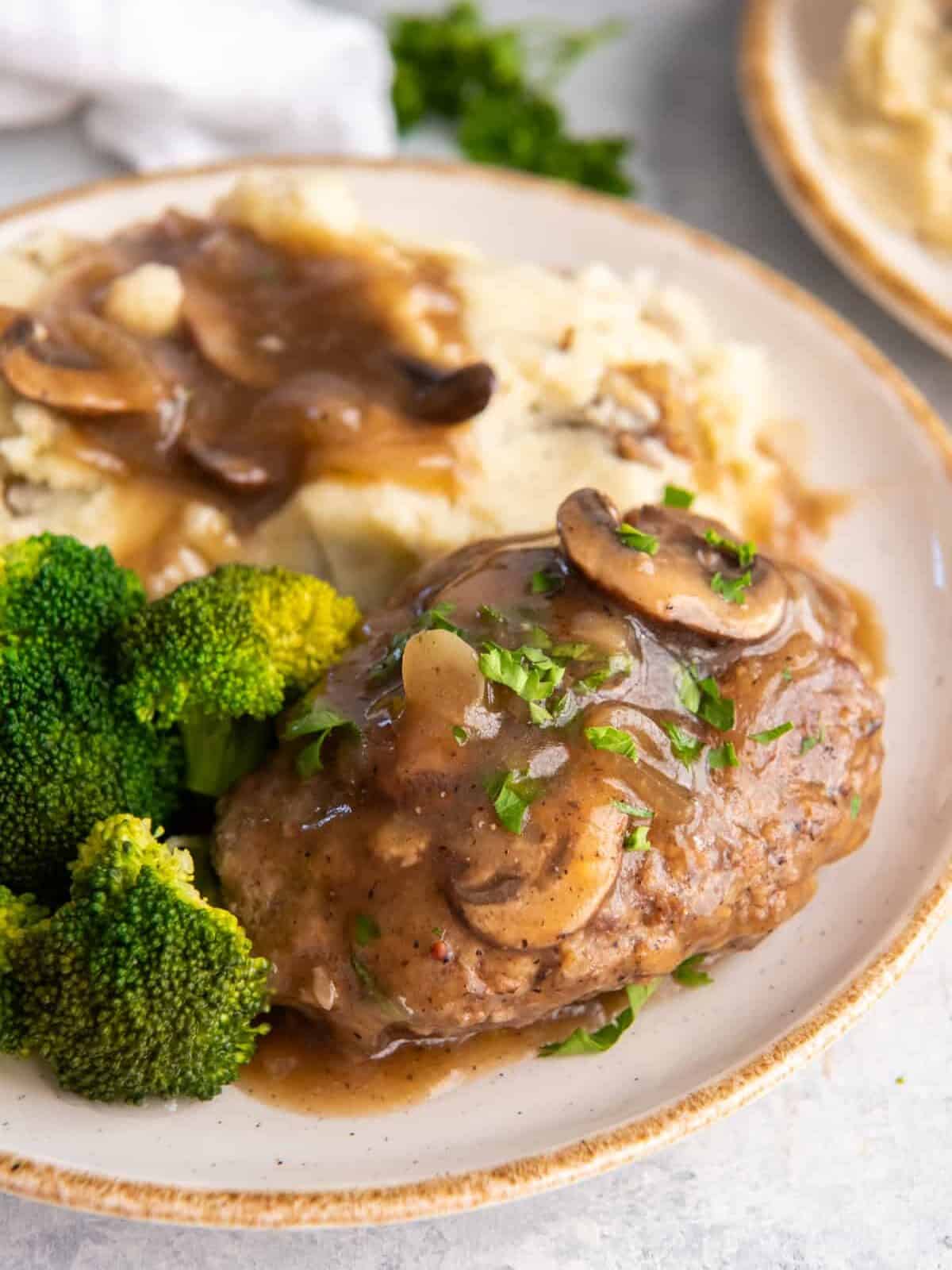 close up of salisbury steak on a white plate with mashed potatoes and broccoli.