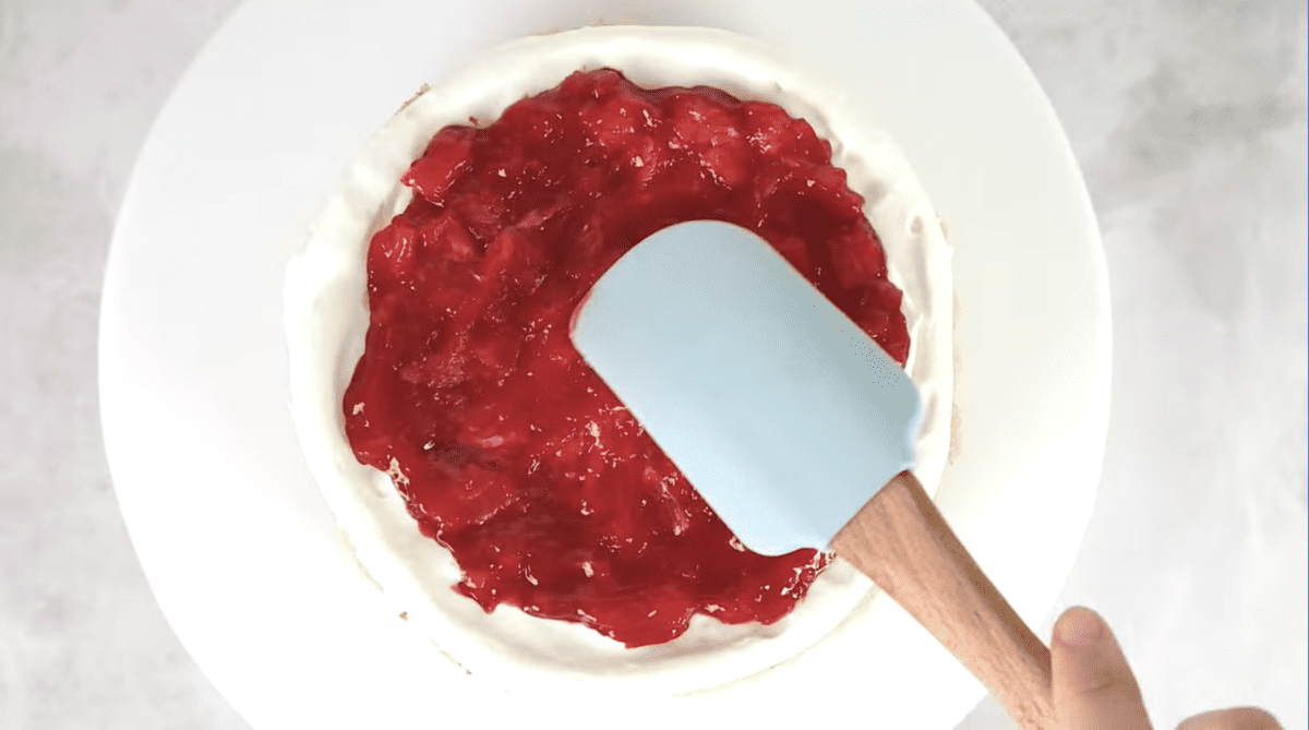 strawberry filling spread over a frosted cake on a cake stand with a rubber spatula.