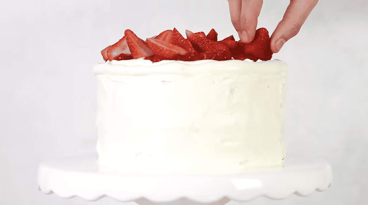 placing a fresh strawberry on top of vanilla cake with strawberry filling on a cake stand.