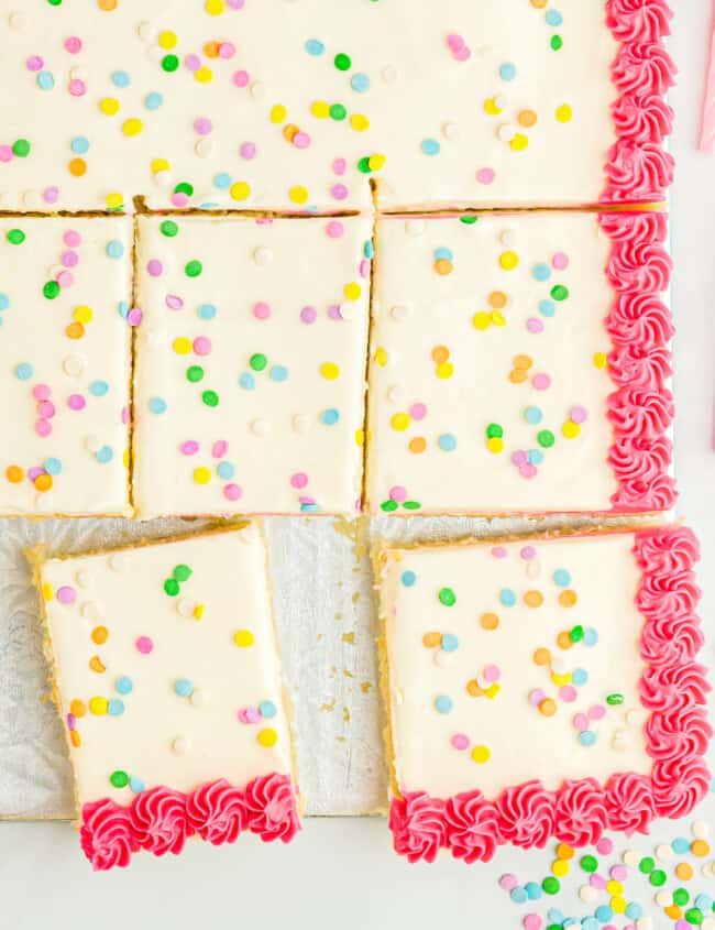 close up overhead view of a partially sliced vanilla cake decorated with white icing with a pink border and sprinkles.