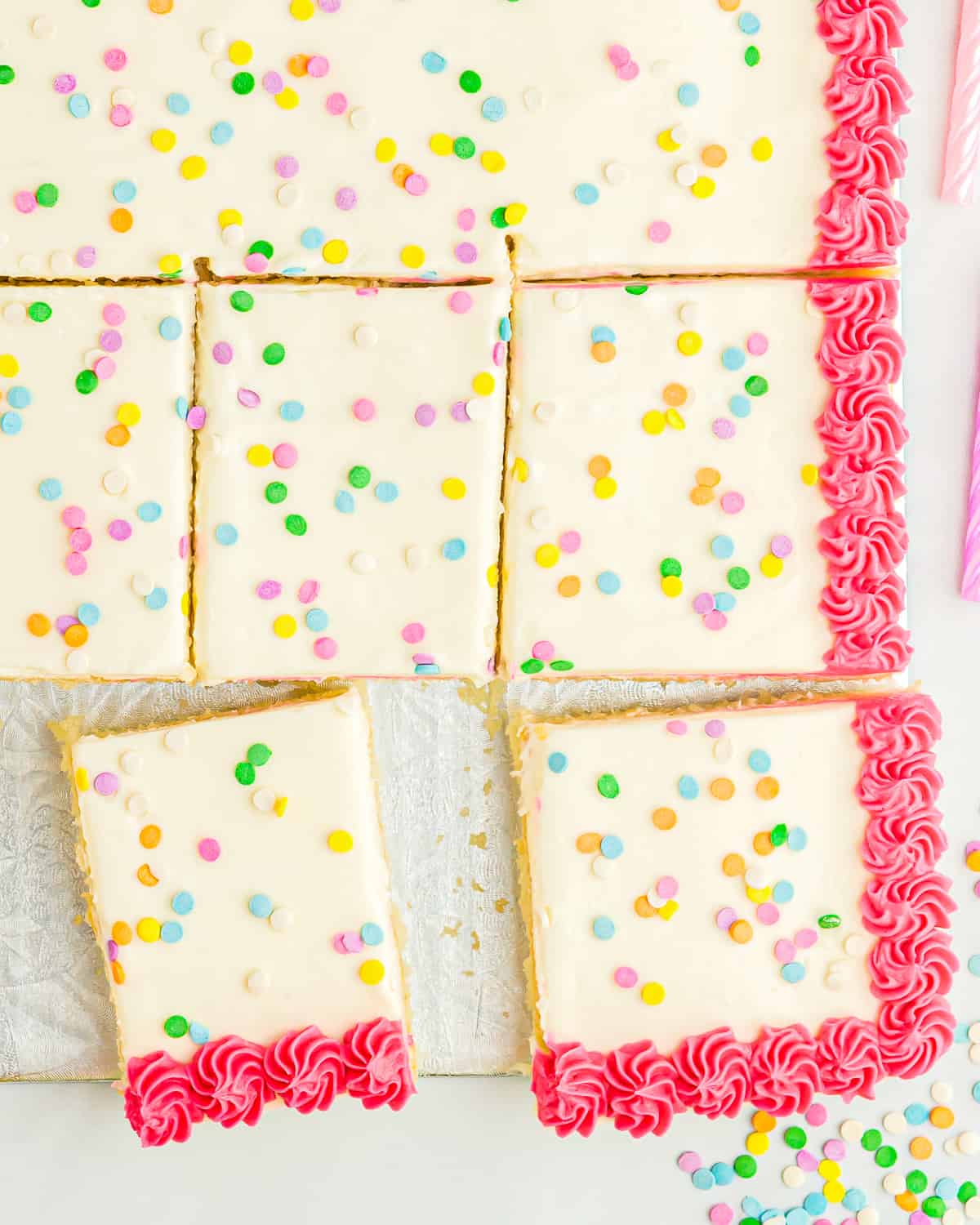 close up overhead view of a partially sliced vanilla sheet cake decorated with white icing with a pink border and sprinkles.